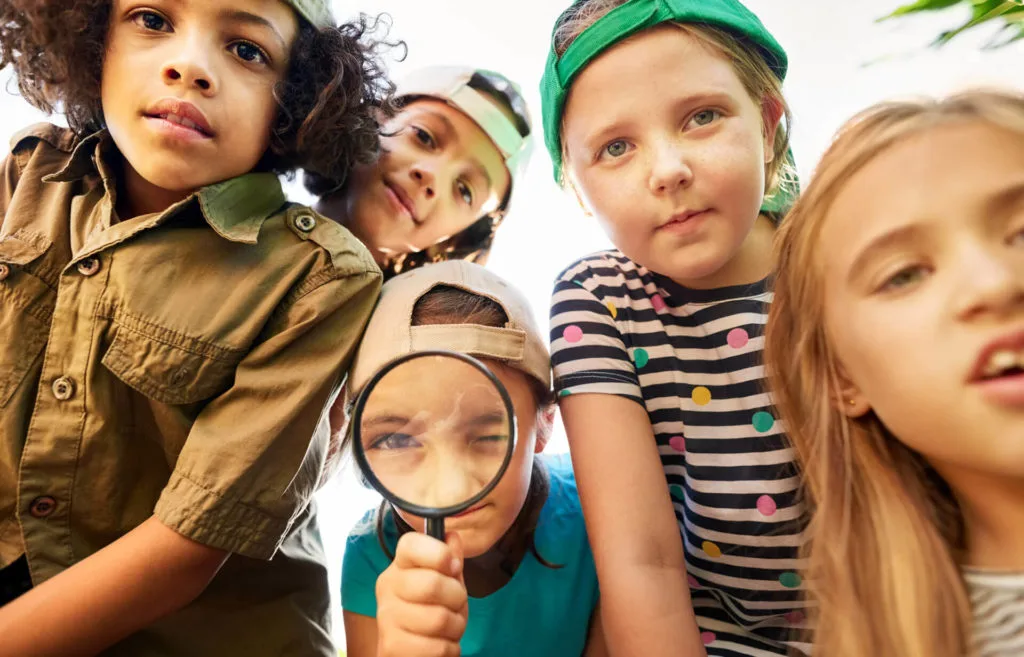 Club der jungen Detektive in Köln: Fünf Kinder beugen sich aus der Froschperspektive über die Kamera; eines von ihnen blickt durch eine Lupe, zwei tragen Basecaps.